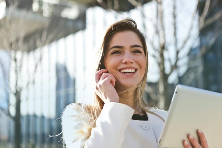 Smiling woman in white coat holding tablet outside, looking confident and professional