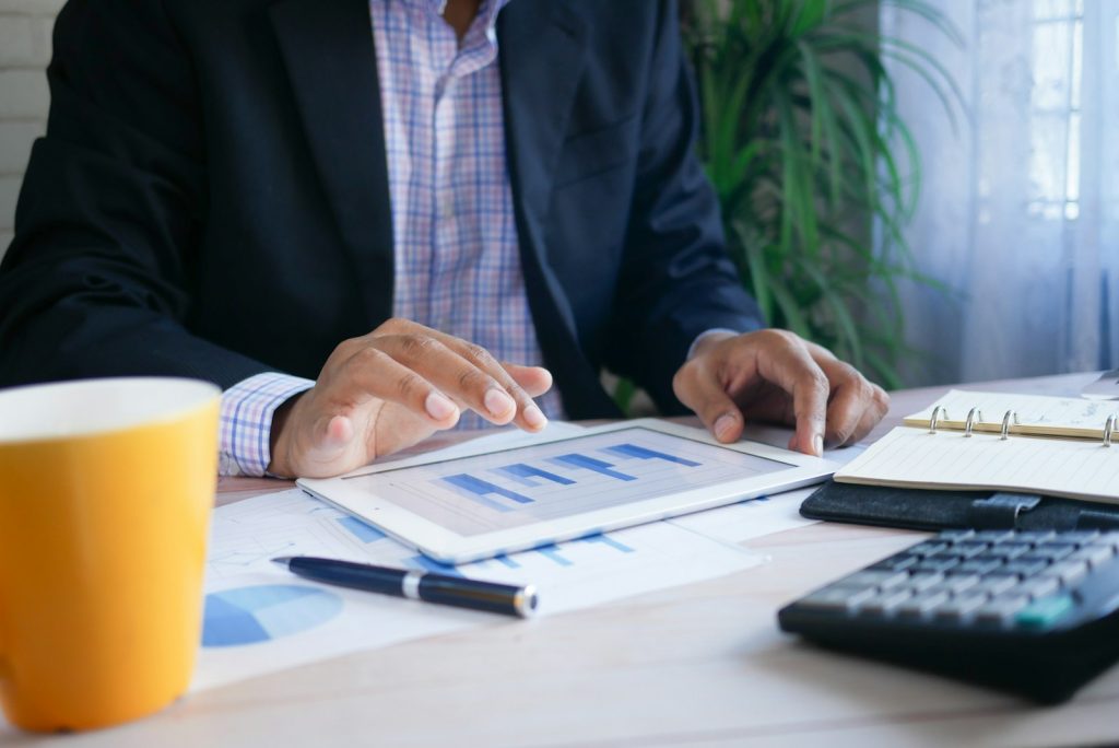 Businessman analyzing data on tablet with charts, surrounded by documents, coffee mug, pen, notebook, calculator