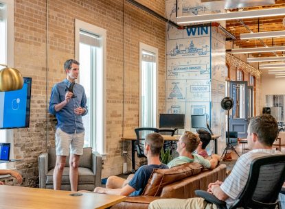 Man giving a presentation to a group in a modern office setting, with a screen showing Proof ALL HANDS 4/24/2019