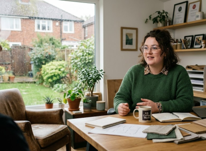 Woman in a cosy home office, sitting at a desk with a coffee mug, surrounded by plants and notebooks